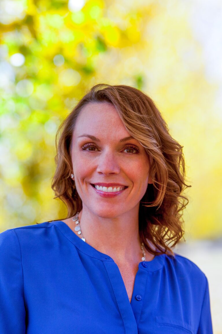 Dr. Rebecca Steinbach wearing a blue blouse with a collar and buttons, along with a round beaded necklace. The blurred background features bright yellow and green colors, suggesting an outdoor setting with foliage.