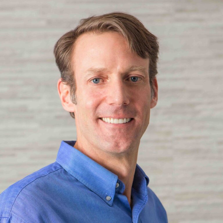 A smiling man with short light brown hair is shown in a professional head-and-shoulders portrait against a neutral background. He is wearing a blue collared shirt, giving a clean and approachable appearance suitable for a professional setting.