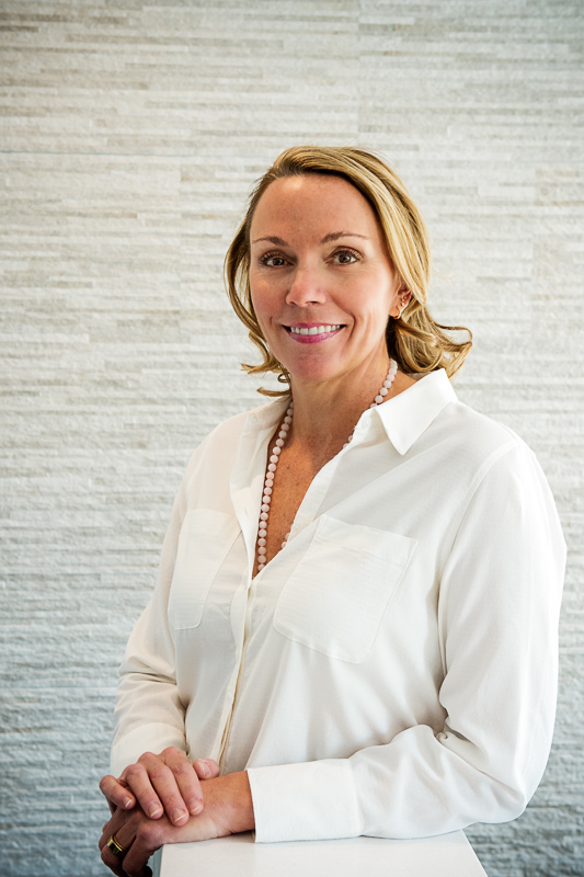 A woman in a white button-up shirt with chest pockets and a pearl necklace, standing against a textured light wall. Their hands are clasped on a white surface.