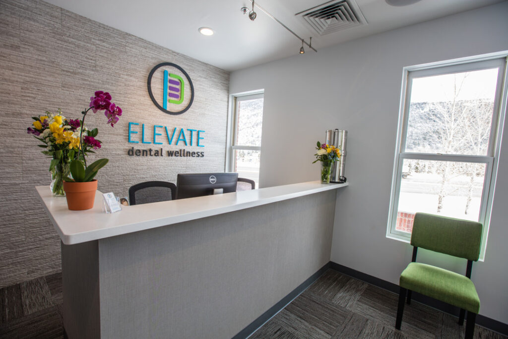 Modern dental reception with white desk, orchid, and logo wall reading "Elevate Dental Wellness"