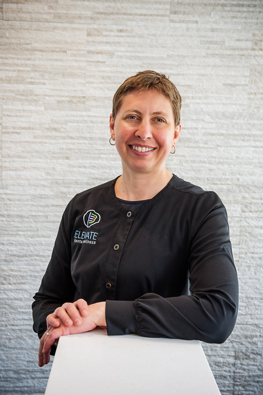 A woman standing behind a counter, wearing a black shirt featuring the logo and text 'ELEVATE dental wellness,' with a light-colored textured wall in the background.