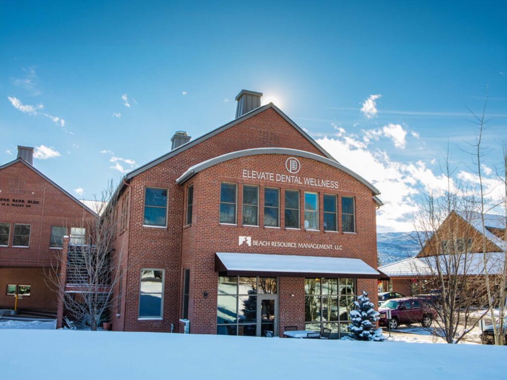 A brick commercial building with the sign “Elevate Dental Wellness” is shown under a clear blue sky, with snow covering the ground and nearby trees. The building has large windows, a curved architectural detail at the roofline, and a professional, welcoming exterior.