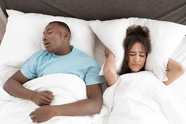 A couple lying in bed, with the man sleeping and snoring loudly, while the woman is awake, looking annoyed and covering her ears with a pillow.