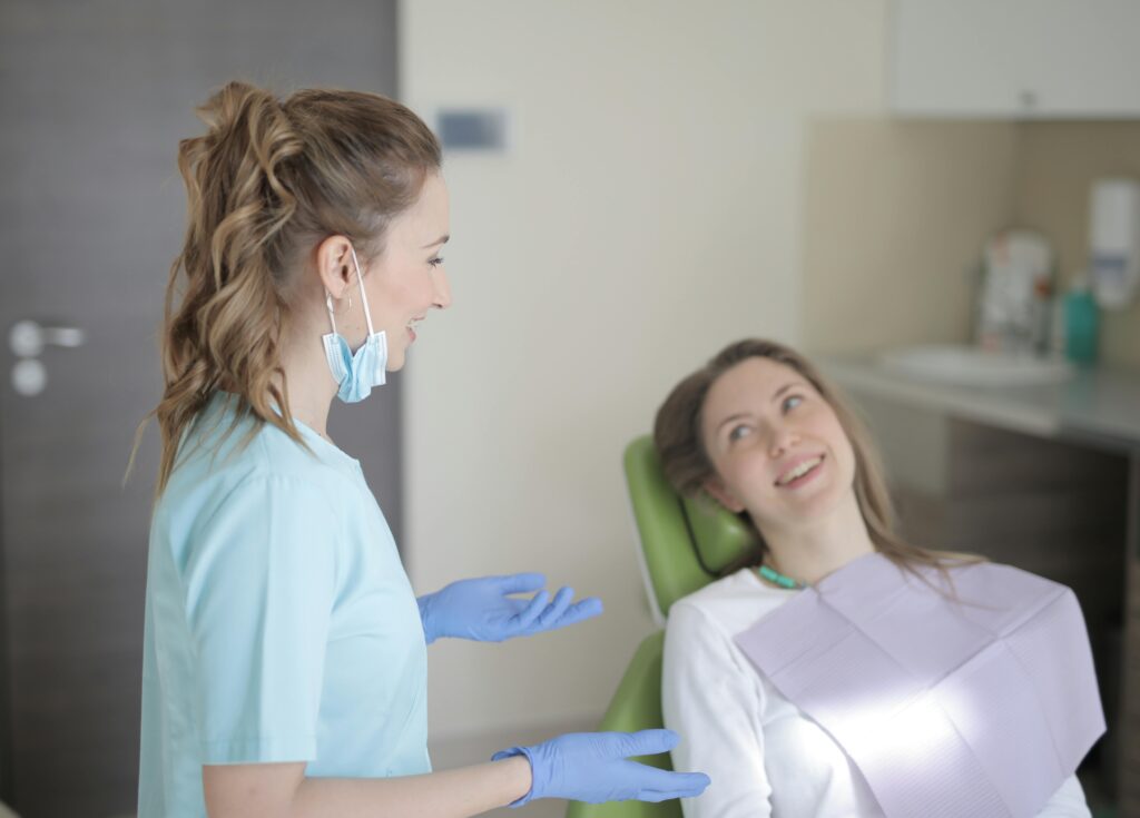 A dentist or dental hygienist wearing a mask and gloves is speaking with a smiling patient who is sitting in a dental chair, indicating a friendly and reassuring interaction before or after a dental procedure.