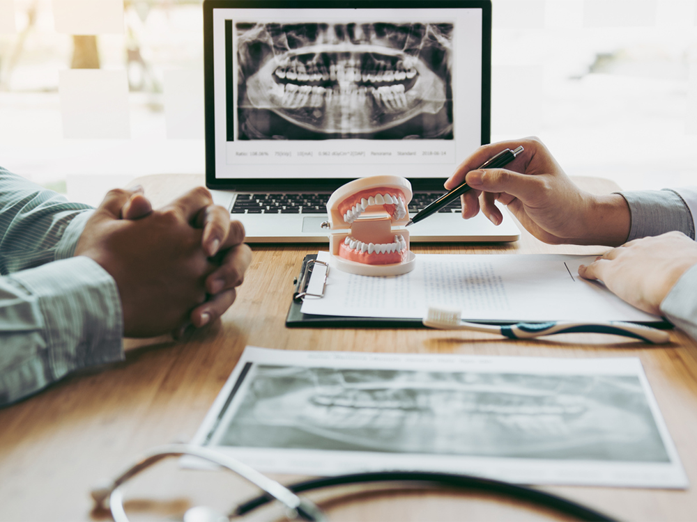 A dental professional explaining a treatment plan to a patient using a dental model and X-ray images displayed on a laptop. The scene includes documents and dental tools on the table, indicating a detailed consultation about the patient's dental health and treatment options.