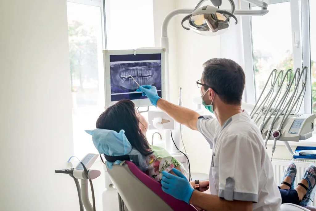 A cheerful young woman with curly hair smiles during a dental consultation with a male dentist in a modern clinic, both wearing masks, creating a friendly and reassuring atmosphere