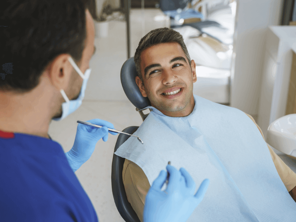 A dentist wearing a mask and gloves is speaking with a smiling male patient who is sitting in a dental chair. The patient looks relaxed and engaged, indicating a positive and reassuring dental visit experience.
