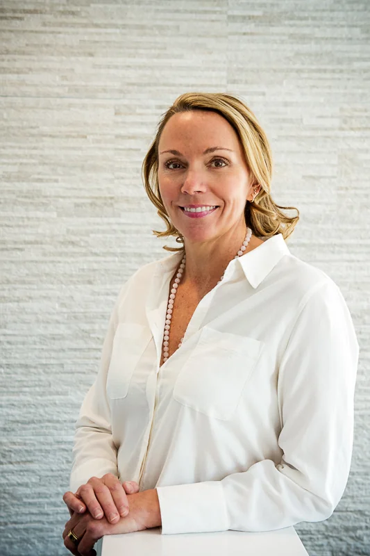 A woman in a white button-up shirt with chest pockets and a pearl necklace, standing against a textured light wall. Their hands are clasped on a white surface.