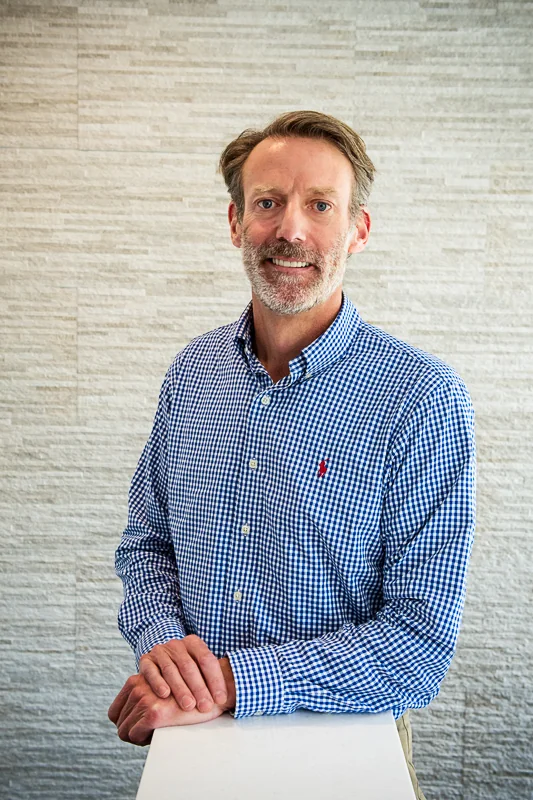 A man wearing a blue and white checkered shirt with a small red logo on the chest, standing in front of a textured wall with hands resting on a white surface.