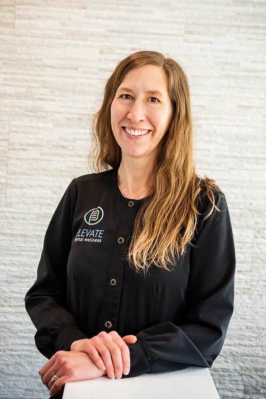 A woman wearing a black button-up shirt featuring the logo of 'ELEVATE Dental Wellness' with a green and purple icon above white text. They are standing in front of a light-colored textured wall, with their hands resting on a white surface.