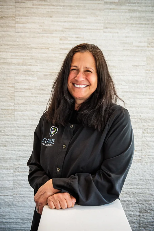 A woman in a black jacket with 'ELEVATE dental wellness' logo, standing in front of a textured light wall, resting hands on a white surface.