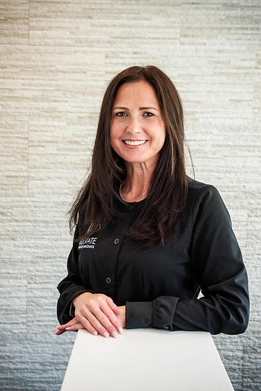 A woman standing behind a counter, wearing a black shirt featuring the logo and text 'ELEVATE dental wellness,' with a light-colored textured wall in the background.