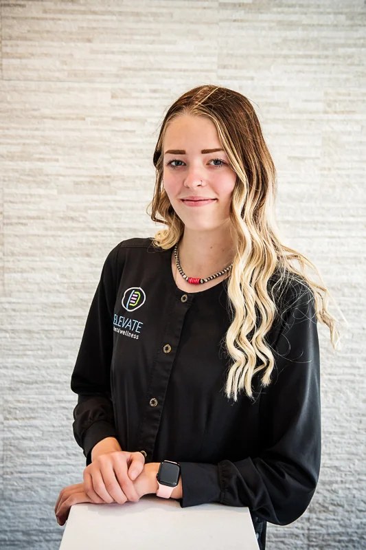 A woman wearing a black shirt with the logo and text 'ELEVATE dental wellness' on the chest, standing in front of a textured light wall. They have long wavy hair with blonde highlights and are accessorized with a necklace and smartwatch.