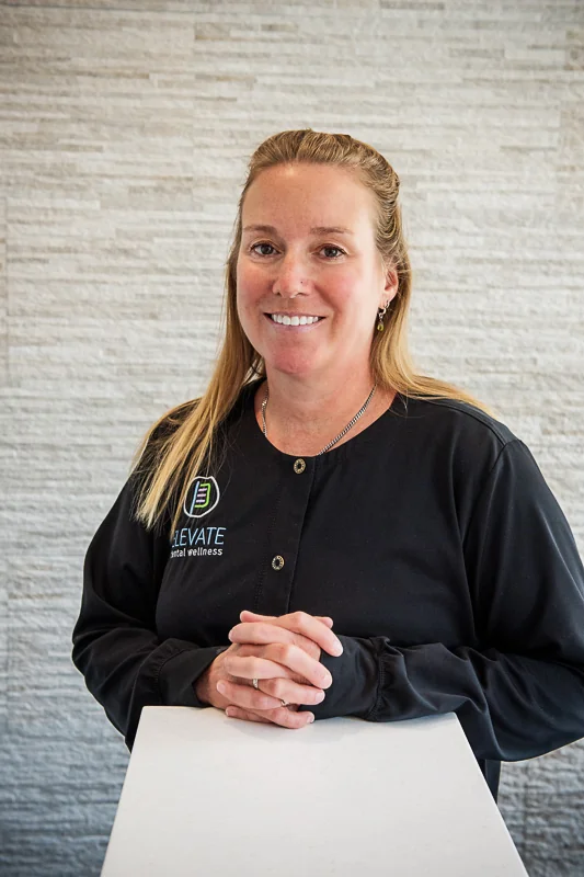 A woman standing behind a counter, wearing a black shirt featuring the logo and text 'ELEVATE dental wellness,' with a light-colored textured wall in the background.