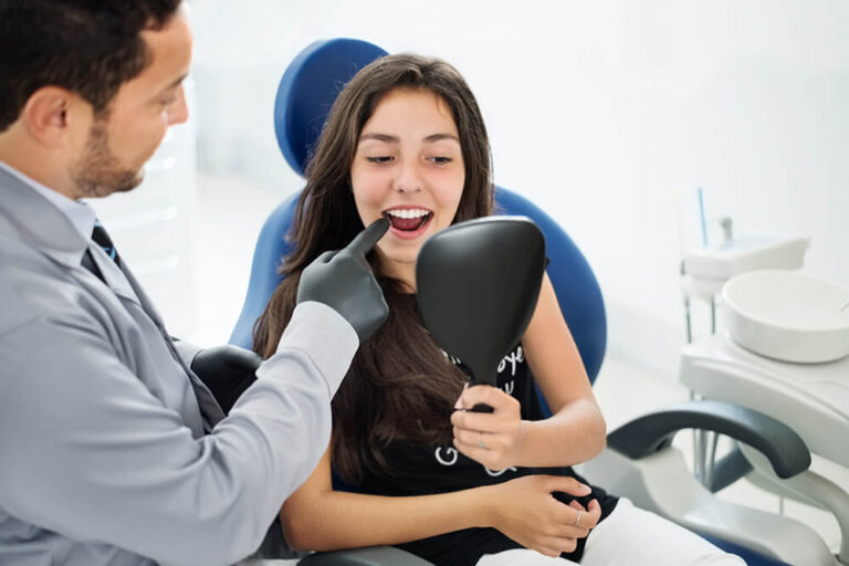 A dental professional wearing gloves examines a patient's teeth during a consultation, with the patient holding a mirror to view their smile. The discussion appears to focus on dental implants as a treatment option for missing teeth.