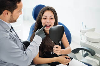 Dentist in gloves pointing at patient’s teeth while patient holds handheld mirror, seated in dental chair with clinic equipment visible.